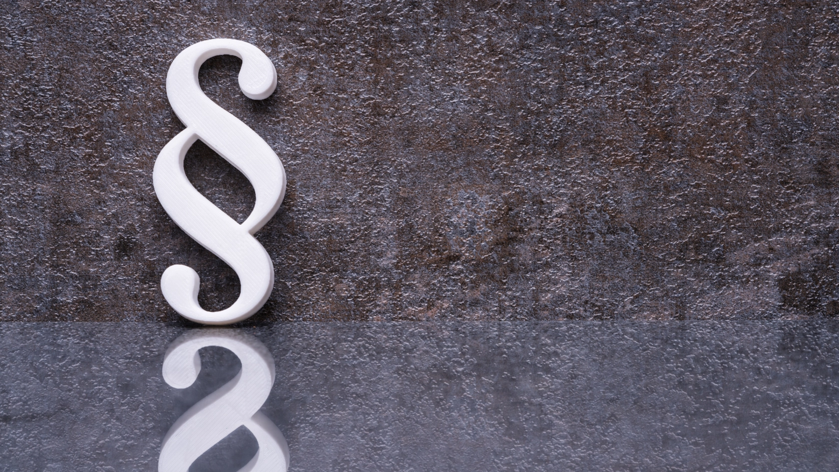Ein weißes Paragraphensymbol steht vor einer rostigen Wand und spiegelt sich im Boden