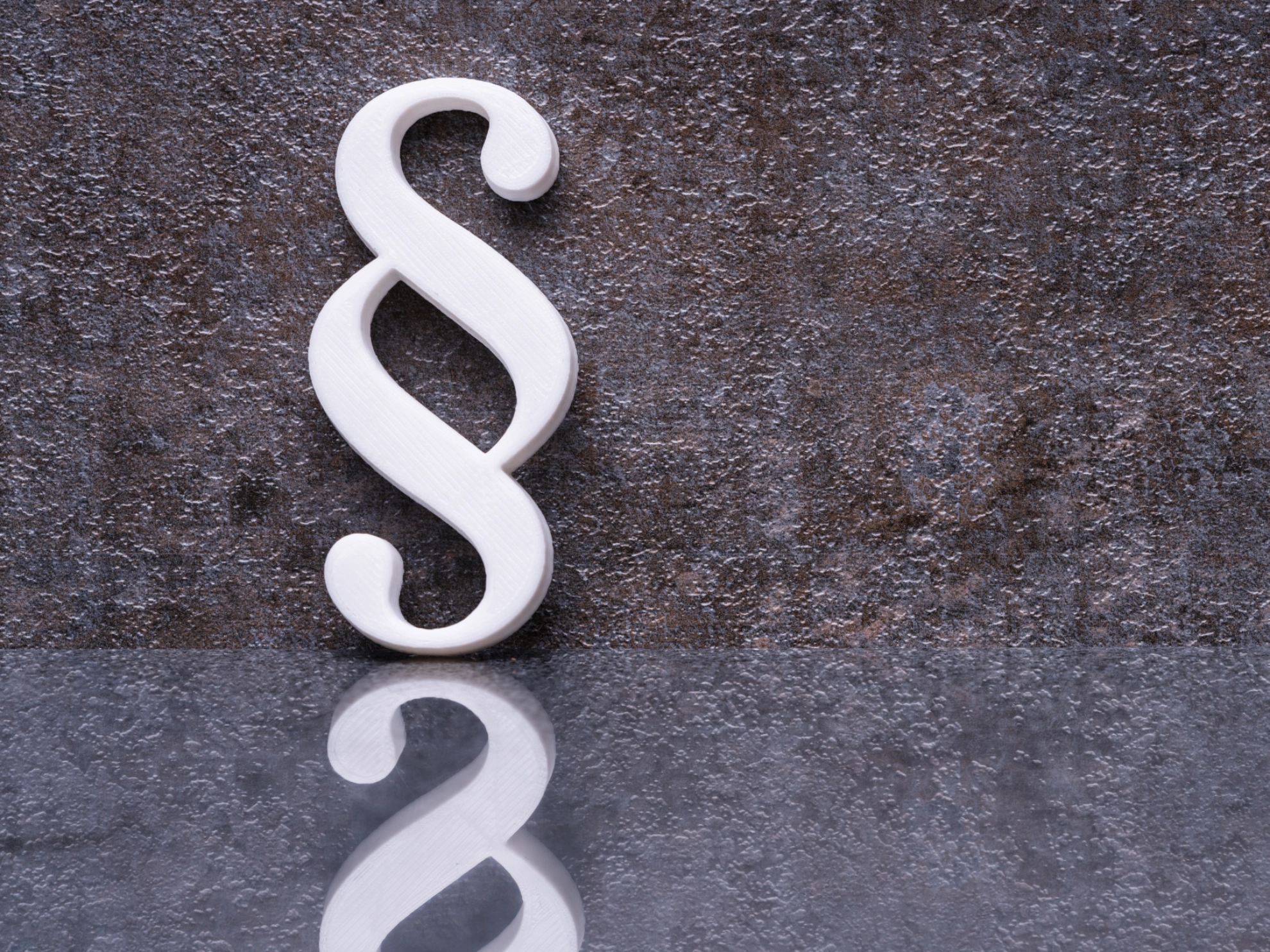 Ein weißes Paragraphensymbol steht vor einer rostigen Wand und spiegelt sich im Boden