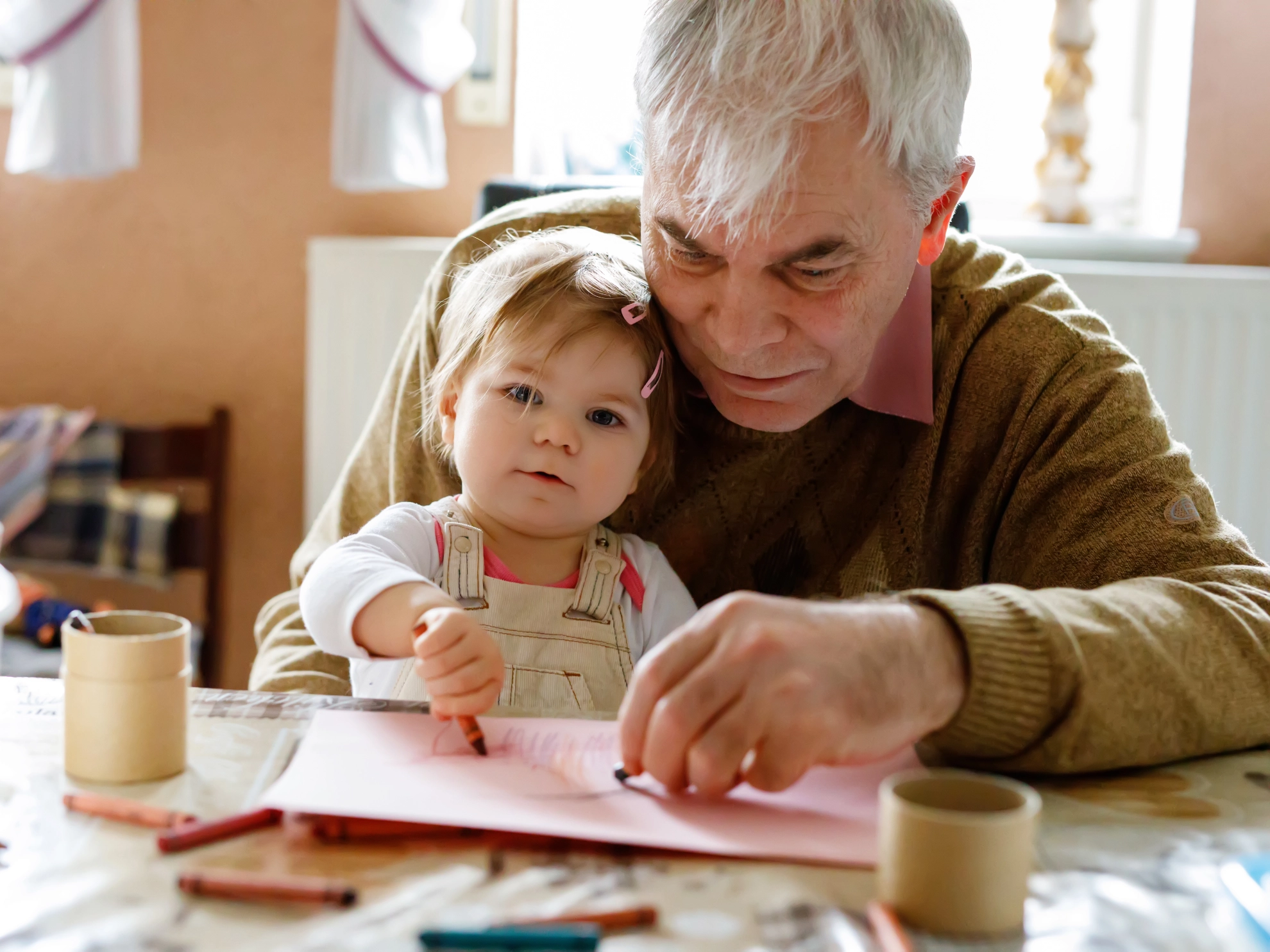 Opa malt mit seiner Enkeltochter ein Bild mit Wachsmalstiften. Sie sitzt dabei auf seinem Schoß.