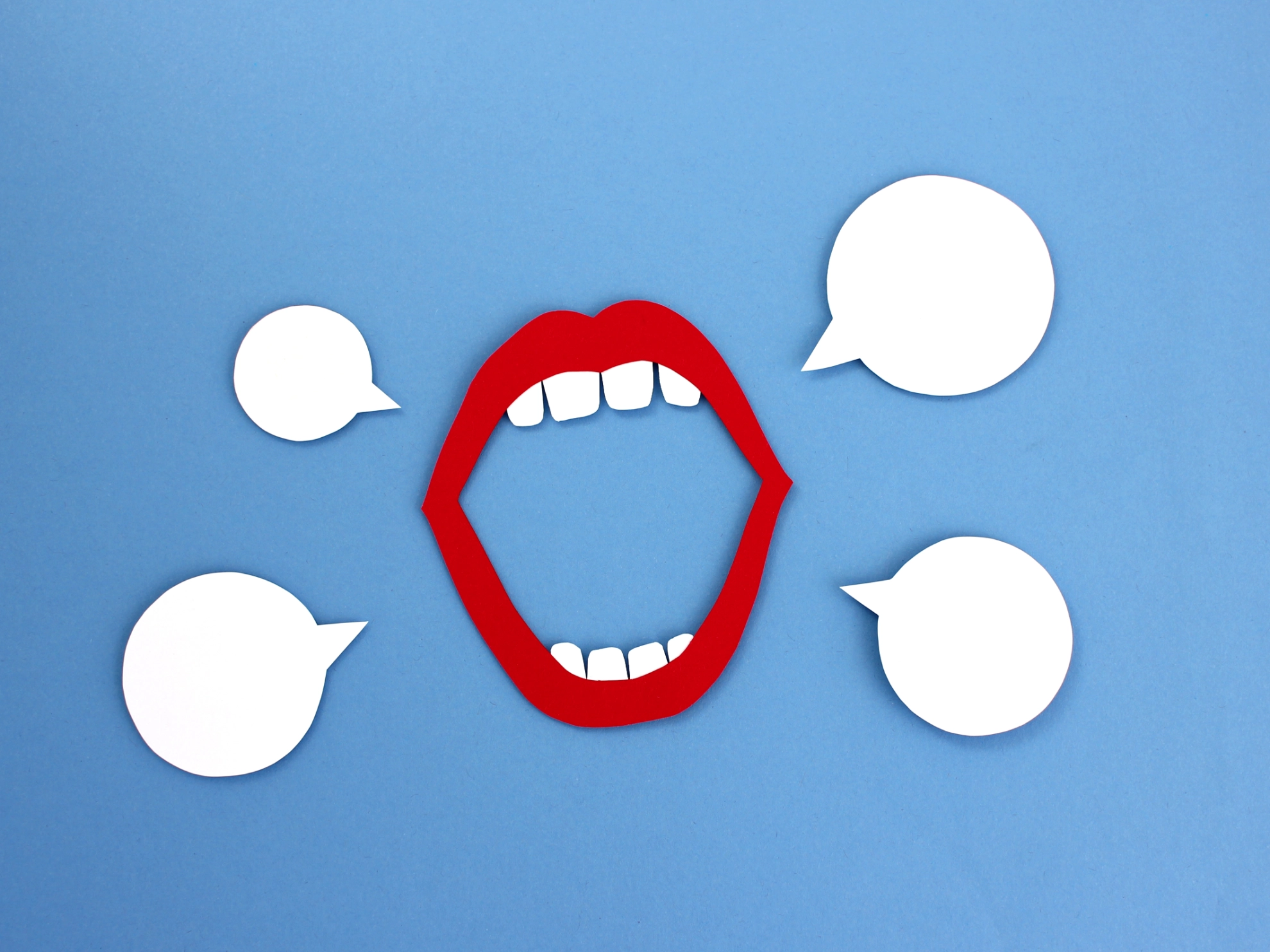 Ein aus Karton ausgeschnittener Mund und vier weiße Sprechblasen vor blauem Hintergrund.